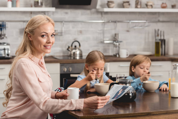 hymyilevä äiti ottaa kupin kahvia, kun istuu keittiössä lasten kanssa aamiaisen aikana ja katselee kameraa
 - Valokuva, kuva