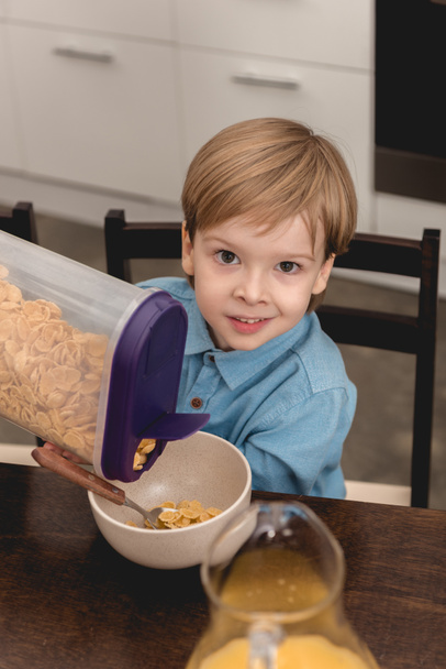 adorable little kid pouring cereal in bowl for breakfast and looking at camera - Foto, Imagem