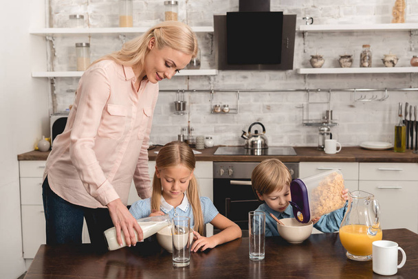 mother pouring milk into cereal for adorable little kids during breakfast - Foto, afbeelding