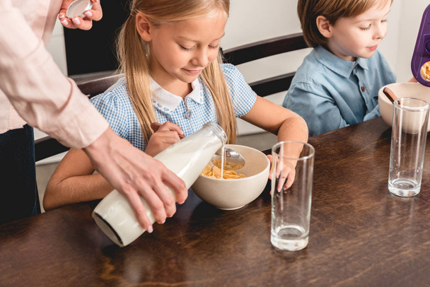 περικοπεί πλάνο της μητέρας ρίχνει το γάλα σε δημητριακών για τα παιδιά κατά τη διάρκεια του πρωινού - Φωτογραφία, εικόνα