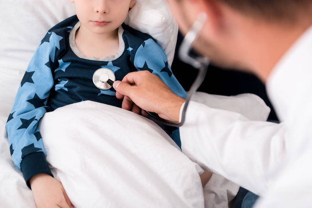 bijgesneden schot van kinderarts litening childs adem met de stethoscoop terwijl hij liggend in bed - Foto, afbeelding