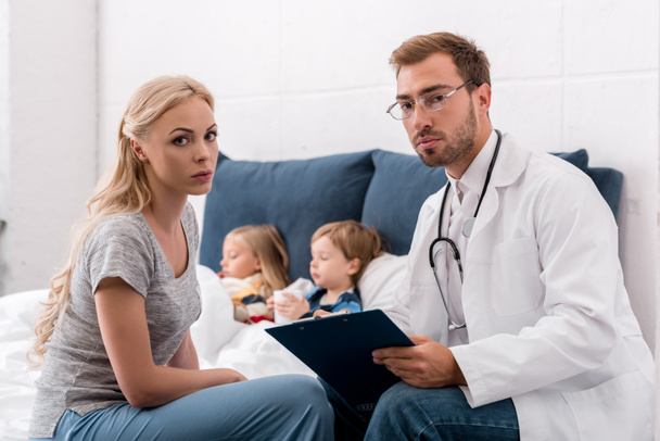 pediatrician and mother looking at camera while sick kids lying in bed on background - Foto, Bild