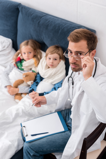 high angle view of handsome pediatrician with clipboard and thermometer looking at camera while sitting near kids lying in bed - Photo, Image