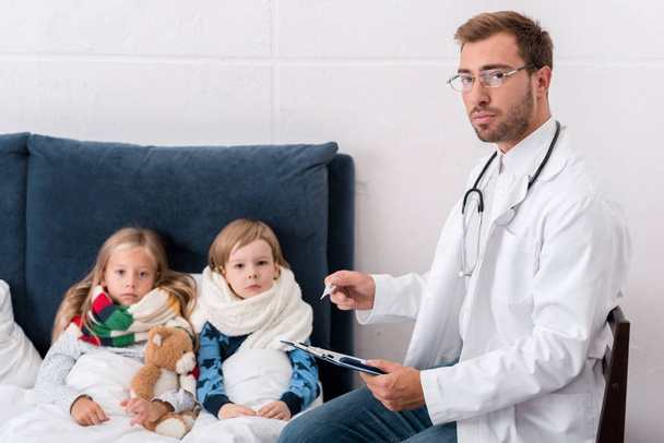 pediatrician writing in clipboard and looking at camera while sitting near kids lying in bed - Foto, immagini