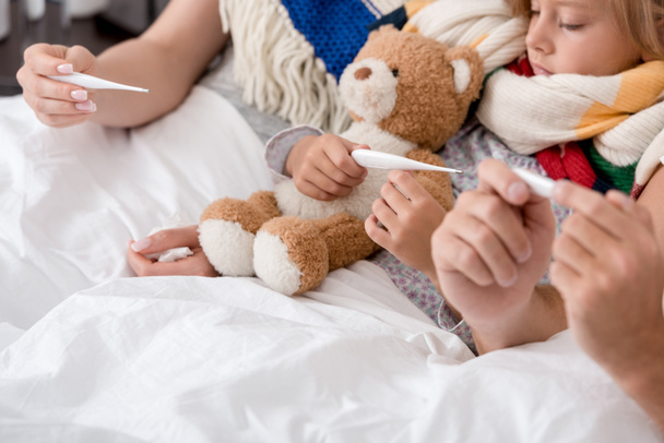 cropped shot of little sick child lying in bed with parents and looking at electric thermometer - Photo, Image