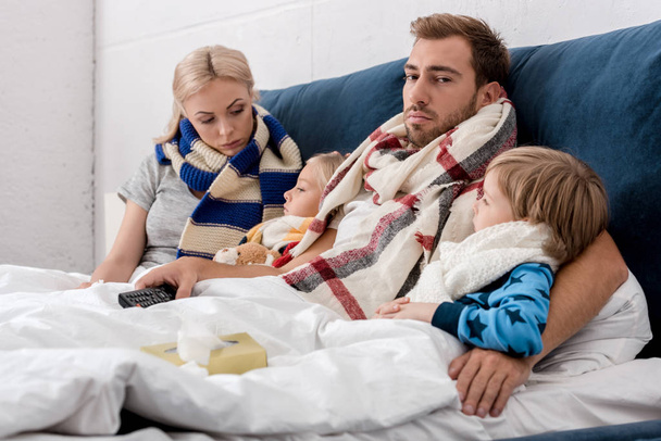 sick young family in scarves lying in bed together with tv remote control - Foto, Imagem