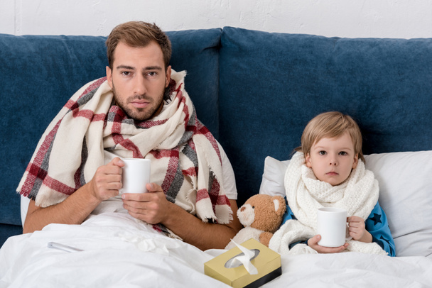 sick father and son in scarves with cups of hot drink sitting in bed and looking at camera - Fotografie, Obrázek