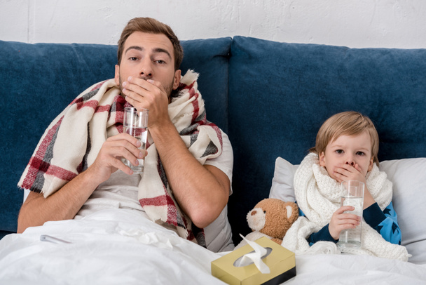 padre e hijo enfermos con vasos de agua tomando pastillas mientras están sentados en la cama y mirando a la cámara
 - Foto, Imagen