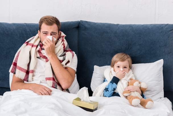 sick father and son blowing noses with paper napkins while lying in bed and looking at camera - Photo, Image
