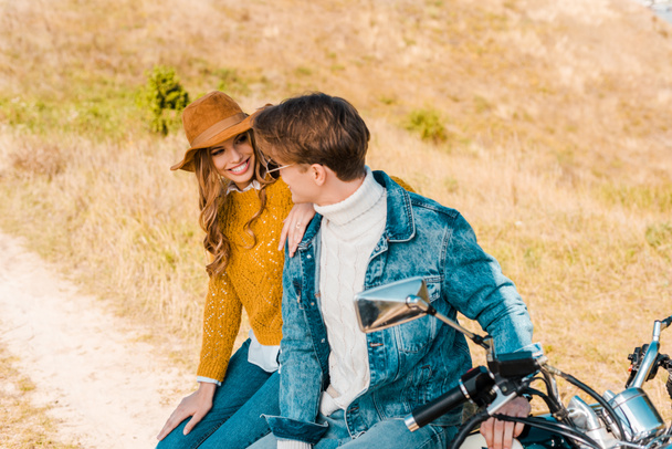 motosiklet üzerinde oturan ve çayır üzerinde birbirine bakarak gülümseyen çift - Fotoğraf, Görsel