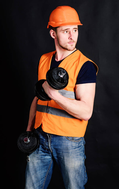 Man in helmet, hard hat holds heavy dumbbells, black background. Strong builder concept. Builder in helmet working out, training. Worker, contractor, builder on strict face with muscular biceps - Foto, imagen