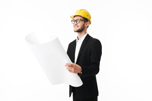 Young smiling bussinesman in eyeglasses and yellow safety helmet happily looking aside holding plan of new project in hands over white background - Foto, Imagem