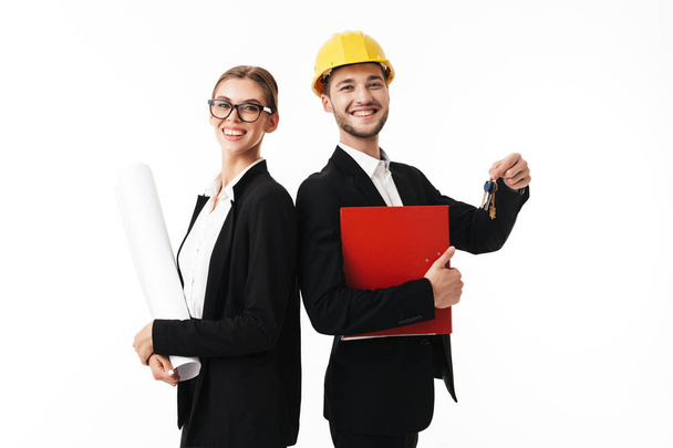 Young smiling business man in yellow safety helmet with folder and keys of new flat in hands and woman in eyeglasses with plan happily looking in camera together over white background - Photo, Image