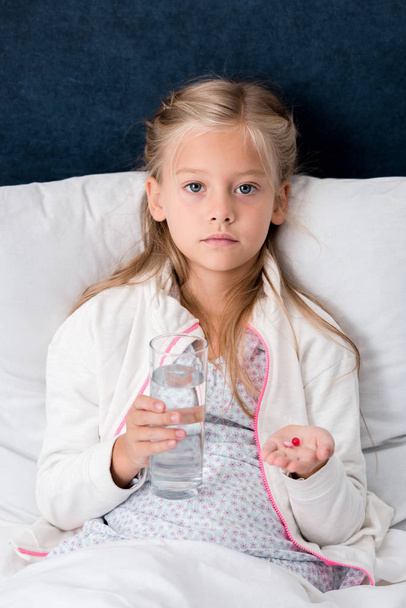 diseased little child with glass of water and pills lying in bed and looking at camera - Photo, image