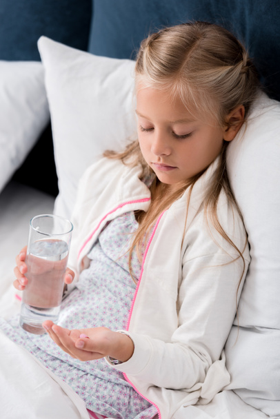 sick little child with glass of water and pills lying in bed - Foto, afbeelding