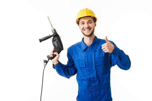 Young smiling foreman in blue uniform and yellow helmet happily showing thumb up gesture looking in camera holding drilling machine in hand over white background - Фото, зображення