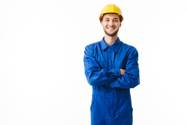 Jonge glimlachend foreman in blauwe uniform en gele helm vrolijk in de camera kijken met handen overtrekken met witte achtergrond - Foto, afbeelding