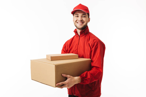 Young cheerful delivery man in red cap and jacket holding parcel in hands happily looking in camera over white background - Photo, Image