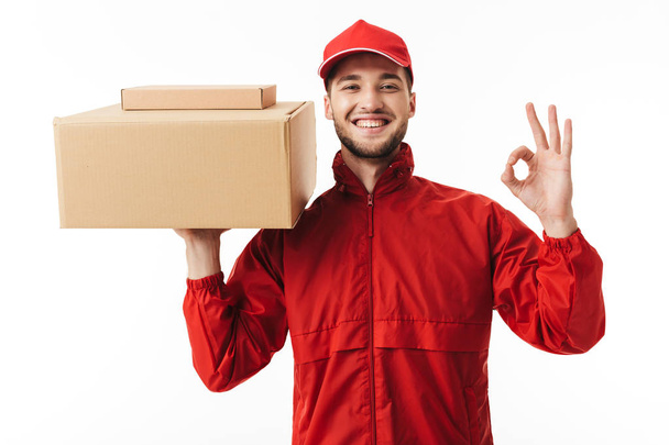 Mladý, usměvavý doručovatel v červené čepici a kabát hospodářství balíčkem v ruce šťastně zobrazeno ok gesto ve fotoaparátu při pohledu přes bílé pozadí - Fotografie, Obrázek