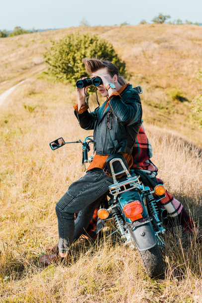 young handsome man with binoculars sitting on vintage motorbike on rural meadow - Foto, Bild