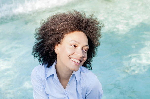 Портрет красивой счастливой афро-американской молодой женщины, улыбаясь смешанной расы женщина расслабляется у фонтана воды, смотрит в сторону, солнечный свет отражается на лице
 - Фото, изображение