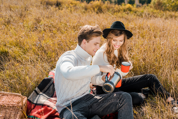 joven sonriente pareja vertiendo bebida caliente del termo en el picnic en el prado rural
   - Foto, imagen