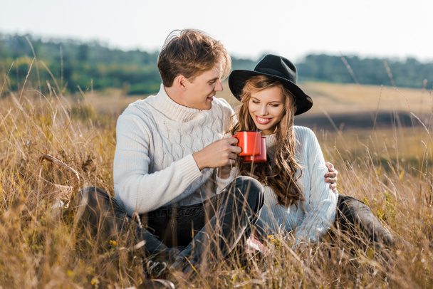 happy young couple with cups relaxing on rural meadow - Φωτογραφία, εικόνα