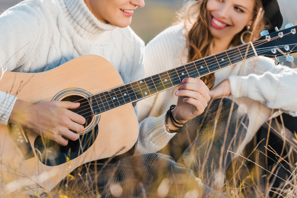 przycięte widzenia uśmiechający się para relaksujący na łące a młody mężczyzna gra na gitarze - Zdjęcie, obraz