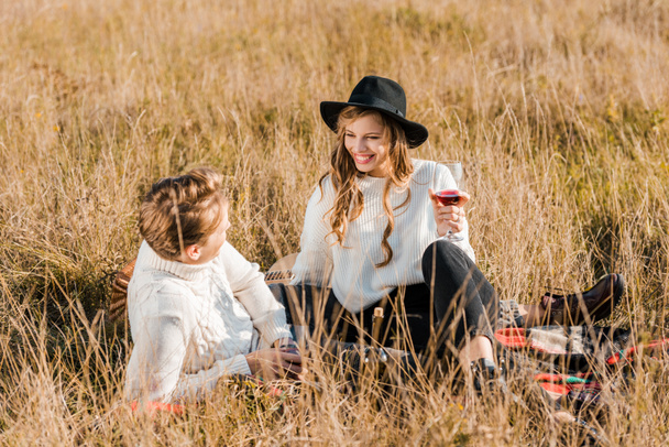 smiling couple holding wineglasses and looking at each other during picnic on meadow - Photo, image