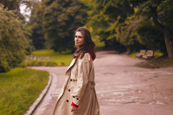 Porträt einer Frau in beigem Trenchcoat und rotem Kapuzenpulli beim Spazierengehen im Park. sie sieht glücklich aus. - Foto, Bild