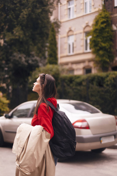 Ritratto di donna che indossa felpa rossa con cappuccio e borsa nera mentre tiene il trench beige nel braccio e gode del paesaggio. passeggiata ragazza turistica in città
. - Foto, immagini