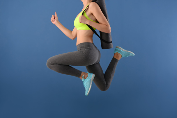 Young woman with yoga mat jumping on color background - Foto, imagen