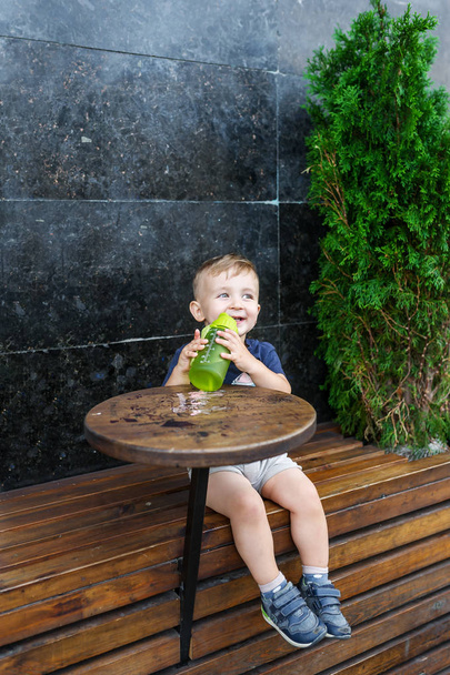 Niño bebiendo de botella de plástico en la calle de la ciudad en la cafetería. Retrato horizontal
 - Foto, Imagen