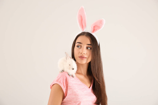 Beautiful young woman with bunny ears and cute rabbit on light background - Zdjęcie, obraz