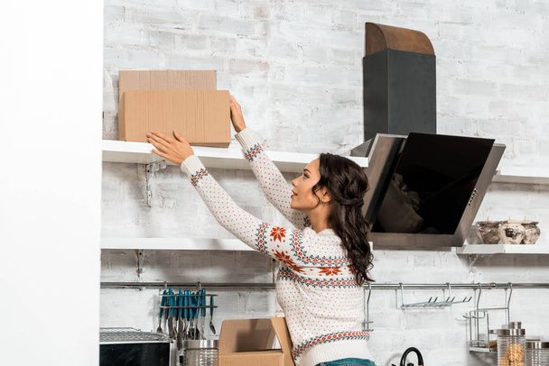 enfoque selectivo de la mujer joven tomando caja de cartón de estante en la cocina en el nuevo hogar
 - Foto, imagen