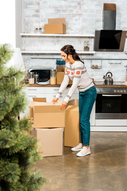 επιλεκτική εστίαση της γυναίκας που στέκεται κοντά σε κουτιά από χαρτόνι στην κουζίνα με Χριστουγεννιάτικο δέντρο κατά τη διάρκεια της μετεγκατάστασης στο νέο σπίτι  - Φωτογραφία, εικόνα