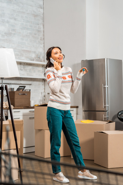 happy young woman talking on smartphone and holding keys in kitchen with cardboard boxes at new home  - Фото, зображення