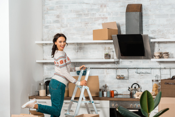 laughing young woman standing on ladder in kitchen with cardboard boxes during relocation at new home  - Photo, Image