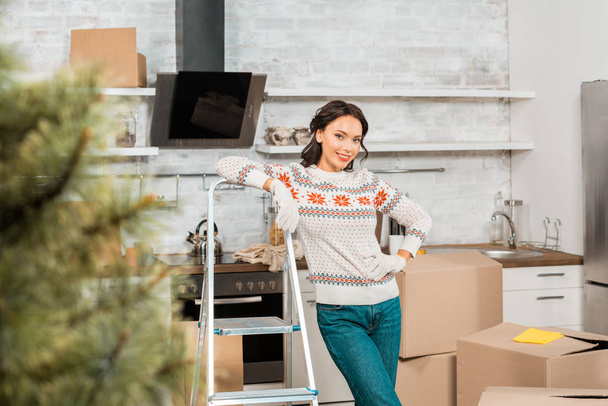 šťastná mladá žena v pracovní rukavice stojící vedle žebříku v kuchyni s kartony v novém domově  - Fotografie, Obrázek