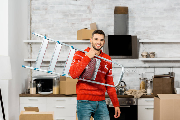 alegre joven que lleva la escalera en la cocina con cajas de cartón durante la reubicación en un nuevo hogar
  - Foto, imagen