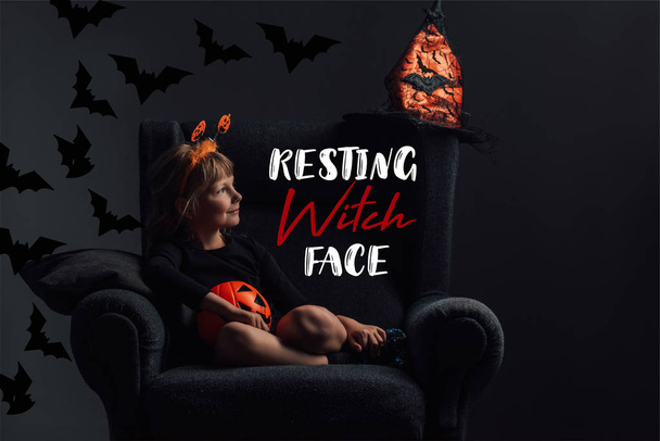 entzückendes Kind im Halloween-Kostüm im Sessel im dunklen Raum mit "ruhendem Hexengesicht" Schriftzug - Foto, Bild