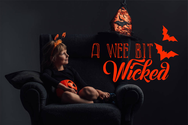 adorable kid w halloween kostium odpoczynku w fotelu w ciemnym pokoju z napisem "wee bit wicked" - Zdjęcie, obraz