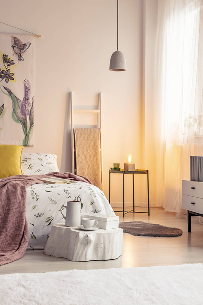 Warm lamp light in a soft colors bedroom interior with a bed dressed in bedding, cushions and blanket. Painted material art on the wall. Real photo. - 写真・画像