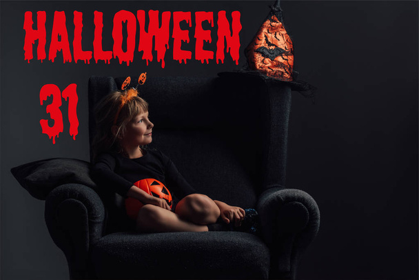 adorable niño en traje de halloween descansando en sillón en habitación oscura con letras de "halloween 31"
 - Foto, Imagen