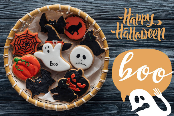 Top view of wicker basket with homemade spooky halloween cookies on wooden table with "happy halloween" and ghost with "Boo" lettering - Photo, Image