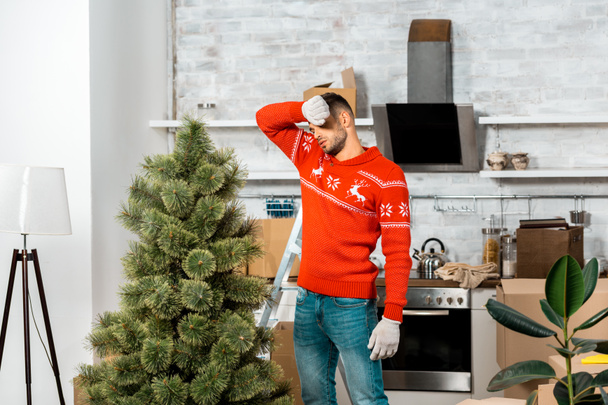 moe man in werkhandschoenen in de buurt van de kerstboom voorhoofd afvegen in keuken thuis - Foto, afbeelding