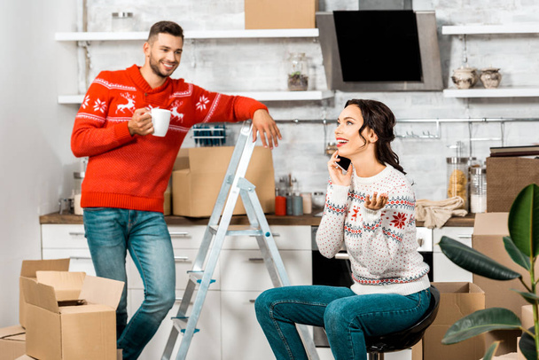 привлекательная счастливая женщина разговаривает по смартфону, пока ее парень пьет кофе на кухне нового дома
  - Фото, изображение
