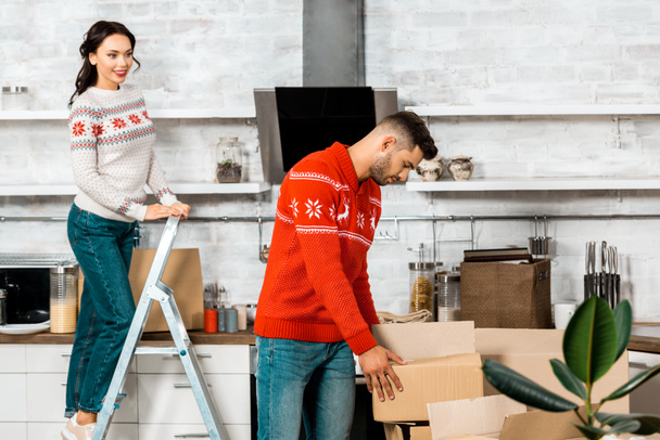 handsome young man taking cardboard box while his girlfriend standing on ladder behind in new home - Photo, Image