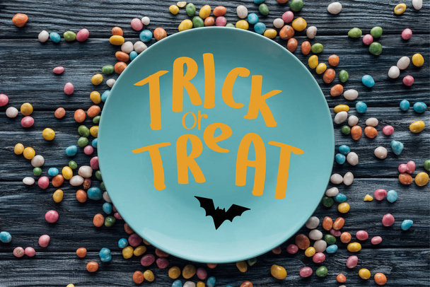 top view of plate with bat and "trick or treat" lettering surrounded by colorful candies on wooden table  - Foto, immagini