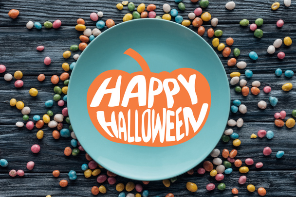 Teller von oben mit Kürbis und "Happy Halloween" -Schriftzug umgeben von bunten Bonbons auf Holztisch  - Foto, Bild